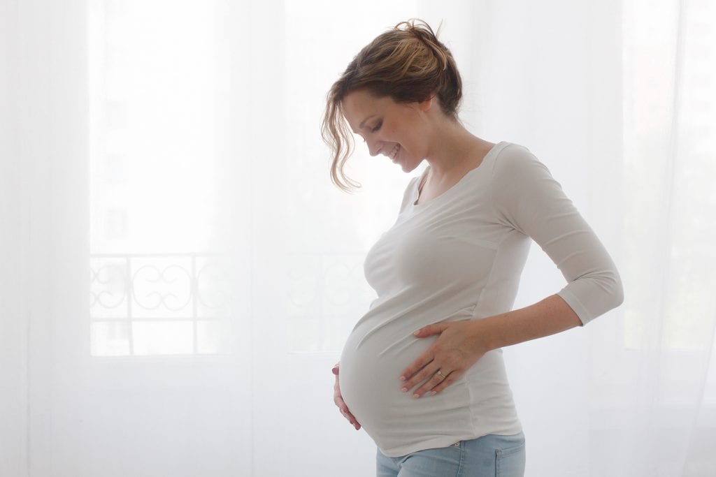 Young pregnant woman with eyes closed embracing her stomach. Horizontal indoors shot.