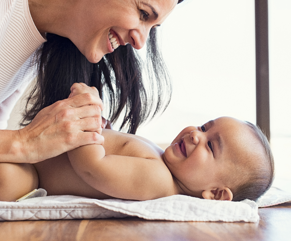 Happy mother playing with baby while changing his diaper. Smiling young woman with baby son on changing table at home. Close up of cheerful mom and toddler boy playing together.
